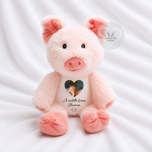 A plush pink pig toy sitting on white fabric, with embroidered pink ears and snout, black bead eyes, and a printed heart-shaped photo on its belly with the text 'A cuddle from Heaven'.