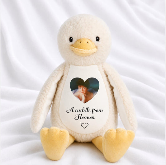 A plush white and yellow stuffed duck toy sitting on white fabric, with a printed heart-shaped photo and the words 'A cuddle from Heaven' on its belly.