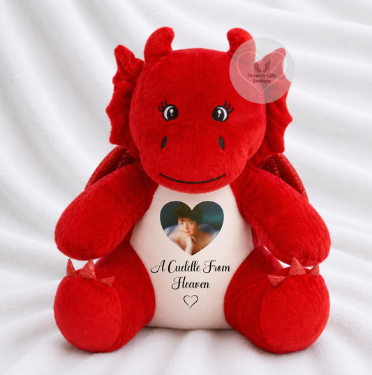 A red plush dragon toy sitting on a white fabric background. The dragon has a white belly patch with a heart-shaped photo cutout and the printed text 'A Cuddle From Heaven' with a small heart below.