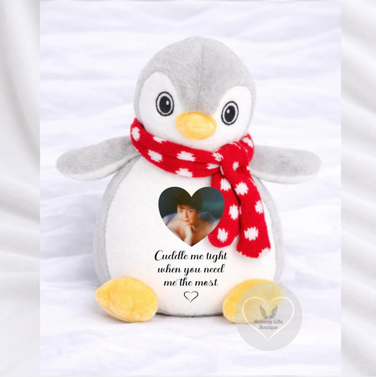 A plush penguin toy with gray and white fur, yellow beak and feet, and a red scarf with white polka dots. The penguin has a heart-shaped cutout on its chest showing a photo, with the text 'Cuddle me tight when you need me the most!' printed below.