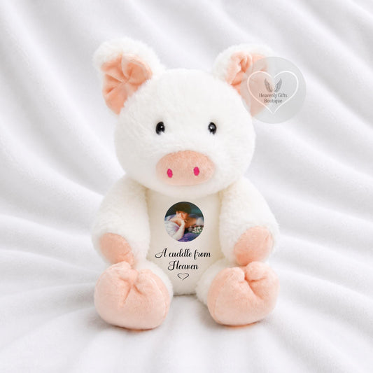 A white plush pig toy with pink ears, snout, and feet, sitting on a white fabric background. The center of the belly has a custom photo cutout with the text 'A cuddle from Heaven' printed below it.
