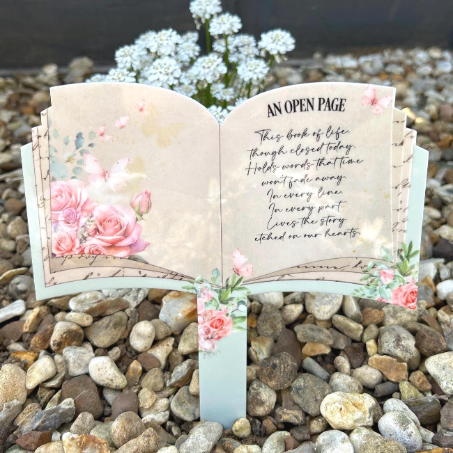 Open book memorial grave marker with pink roses and butterflies displayed outdoors in a remembrance garden