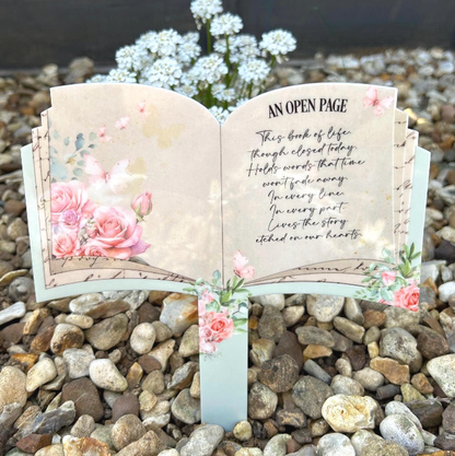 Open book memorial grave marker with pink roses and butterflies displayed outdoors in a remembrance garden