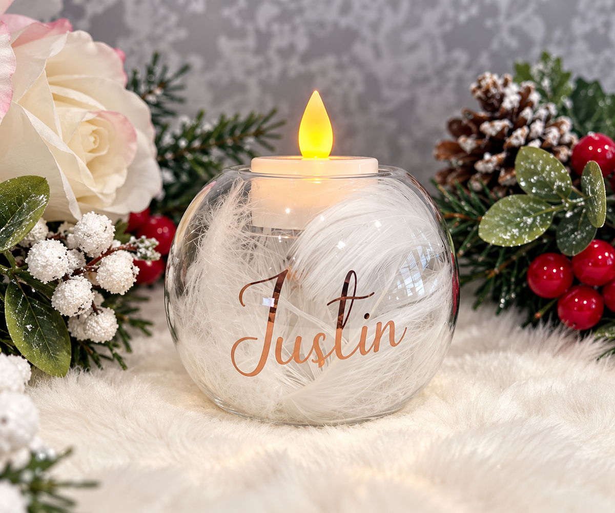 Personalised glass memorial tealight holder filled with white feathers, rose gold Justin name and glowing LED candle among festive foliage